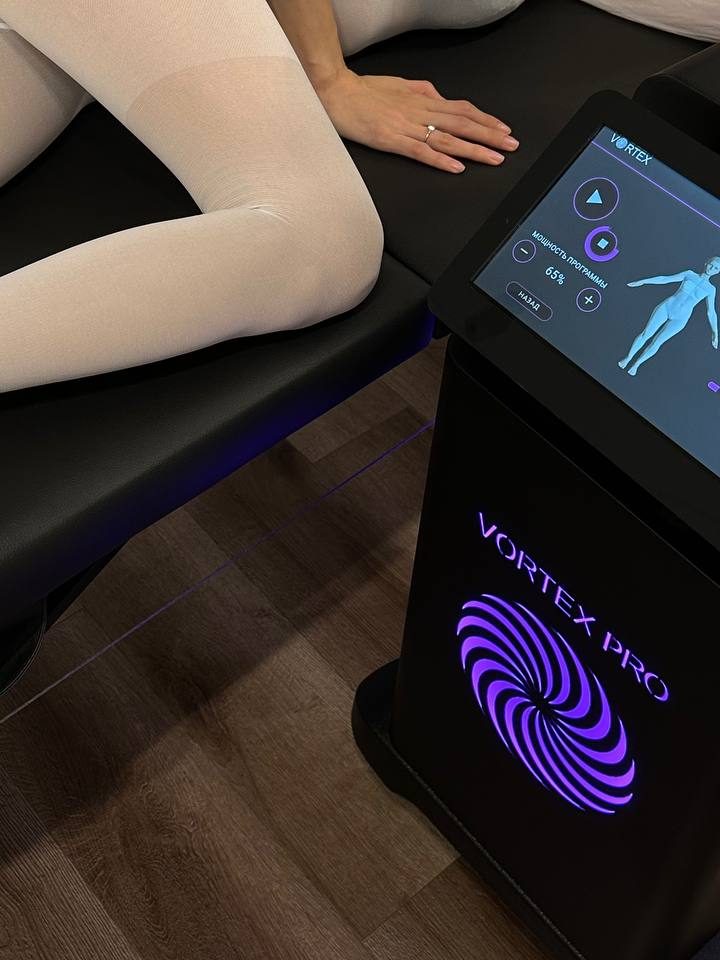 Сеанс LPG: клиентка в белом костюме лежит на боку, рядом аппарат Vortex Pro, мощность программы 65 %
